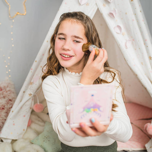 A young girl with long brown hair sits inside a white fabric teepee tent decorated with stars and hearts. She is wearing a white long-sleeve top and a dark green skirt, holding a pink deer-themed compact mirror while applying gold glitter makeup with a brush.