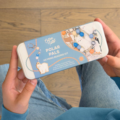 A person holding a metal tin bead keyring kit labeled 'Polar Pals Keyring Beading Kit' over a wooden floor and blue jeans.