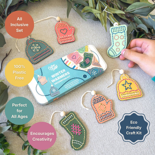 A craft kit showing a tin of winter warmer ornaments surrounded by completed mitten and stocking ornaments, with a hand holding one stocking ornament. Text callouts describe the set as all inclusive, plastic free, 100% plastic free, perfect for all ages, and eco friendly.