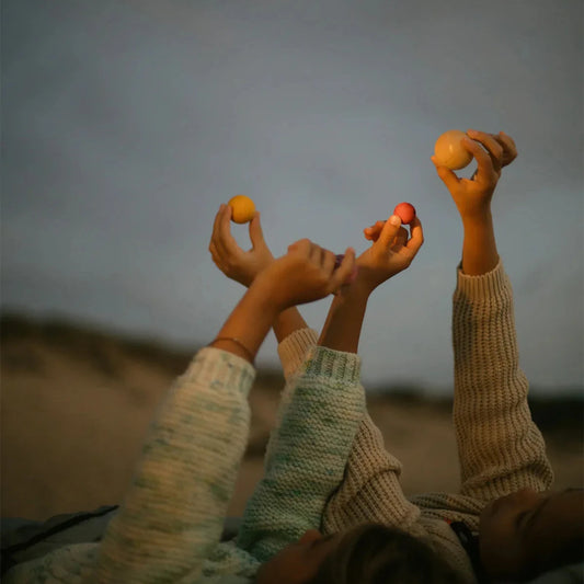 Three children lying on sand at dusk, reaching upward with small wooden planets from Grapat in their hands. They are wearing chunky knit sweaters in cream, light green, and beige tones against a dark, moody sky.