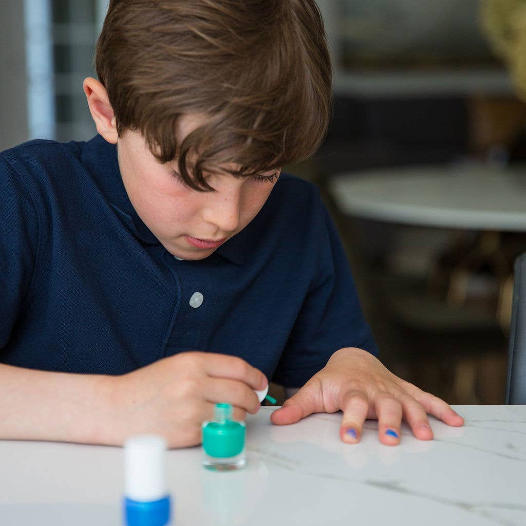 A young boy with brown hair wearing a navy blue polo shirt is sitting at a white marble table, painting his fingernails with a turquoise nail polish. A small bottle of blue nail polish is visible in the foreground.