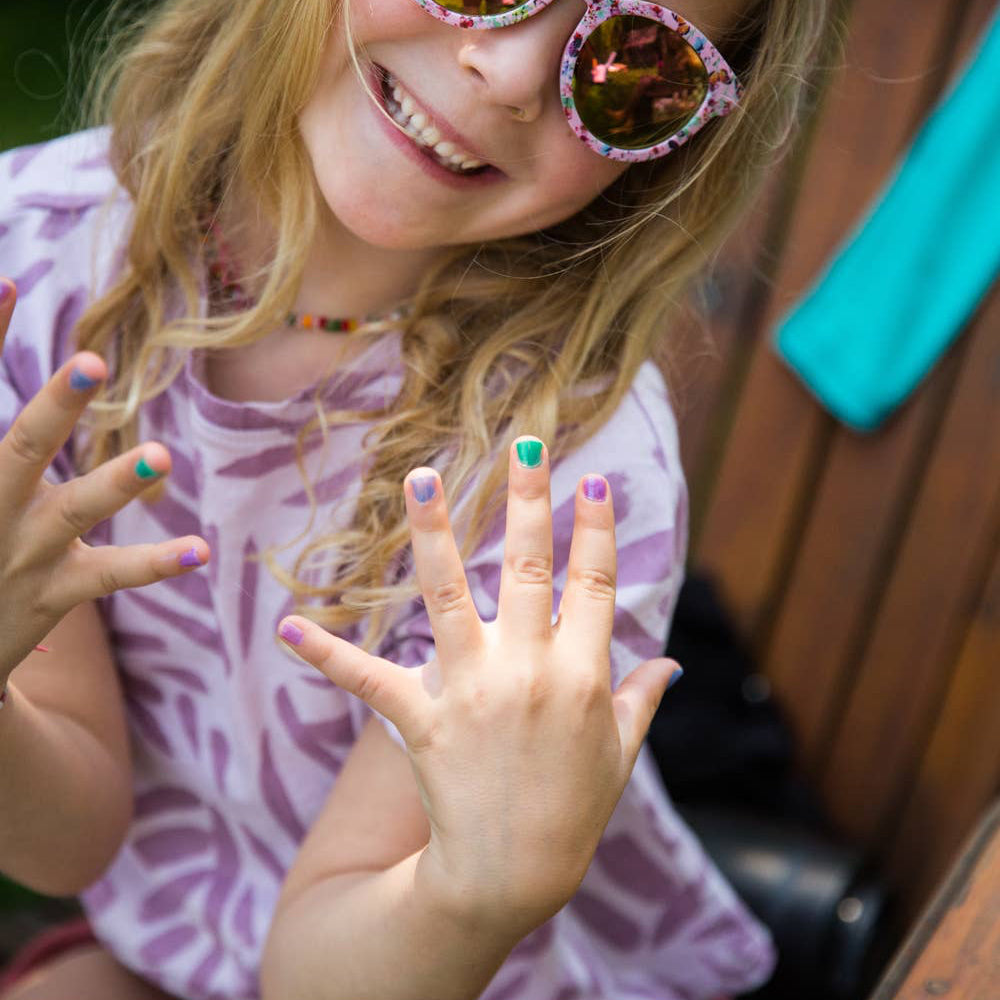 A young girl with blonde hair wearing pink patterned sunglasses and a purple patterned t-shirt, smiling while showing off her colorful painted fingernails.