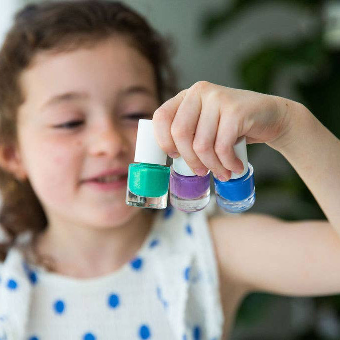 A young girl with brown hair holds up three small Namaki nail polish bottles with white caps. The bottles contain teal green, purple, and blue nail polish, shown against a soft-focus indoor background.