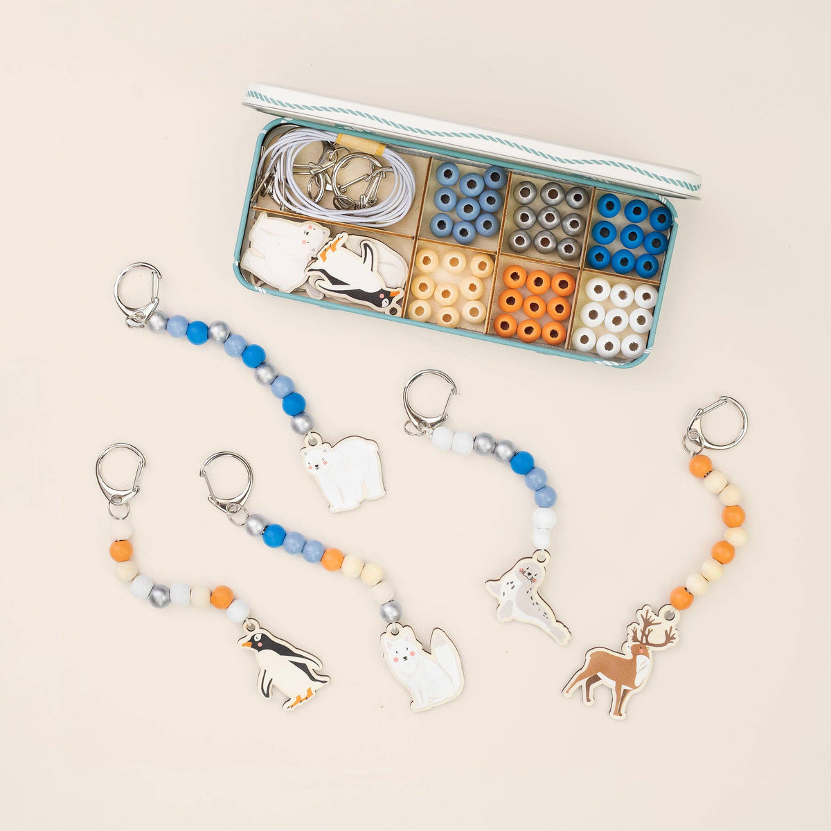 A set of beaded keychains with animal charms (penguin, cat, fox, reindeer) displayed next to an open storage box containing beads, metal rings, and tools on a light beige background.