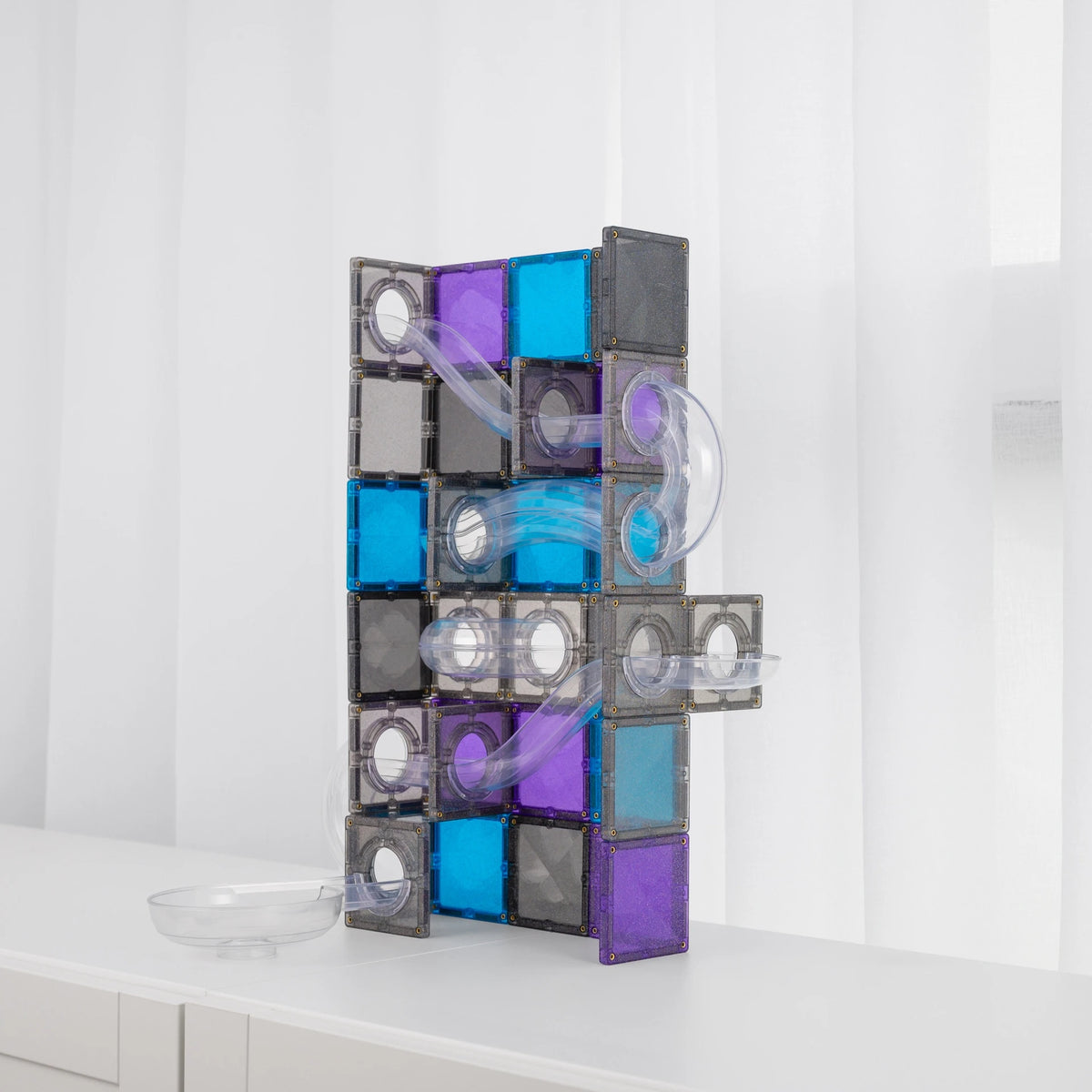 A CONNETIX magnetic tile structure made of translucent gray, purple, and blue square tiles with clear curved tubes, standing on a white surface against a white curtain background.