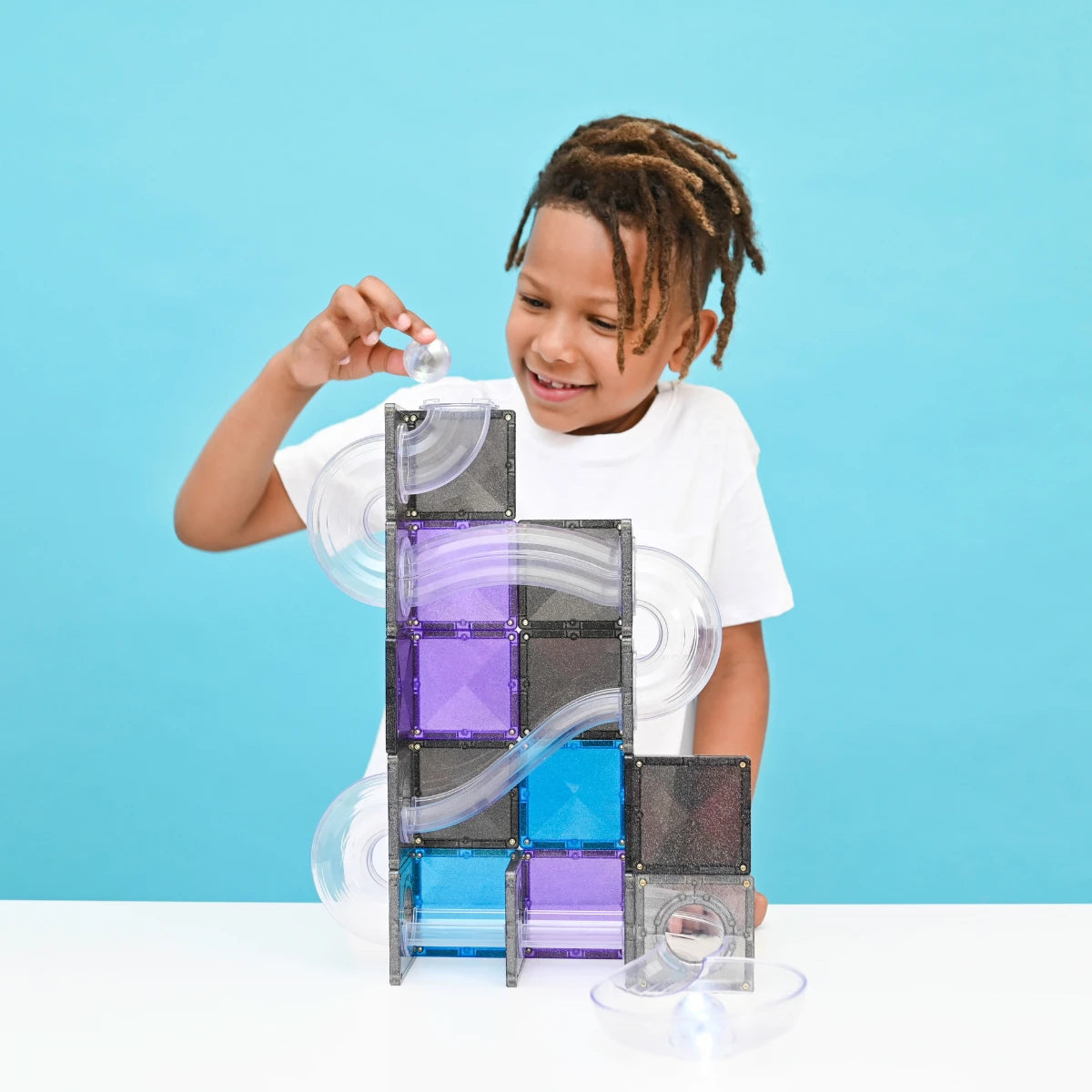 A young boy smiling and building a clear magnetic marble run tower with translucent blocks and purple, blue, and gray panels on a white table against a bright blue background.
