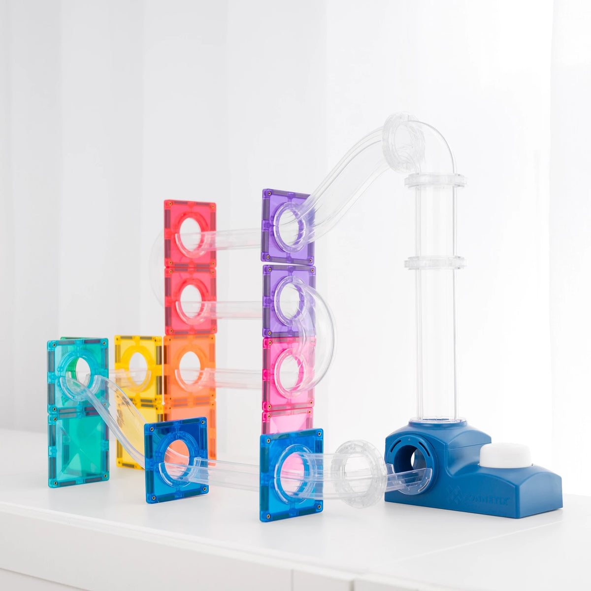 A colorful transparent magnetic tile marble run toy set with multiple acrylic blocks in red, orange, yellow, purple, pink, and blue, connected by clear curved tubes to a blue base on a white surface against a bright white background.