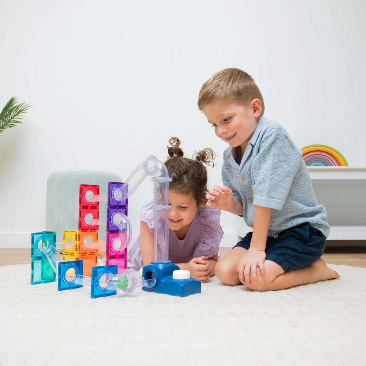 Two children playing with colorful translucent marble-run style magnetic tiles  and tubes on a white rug in a bright indoor room.