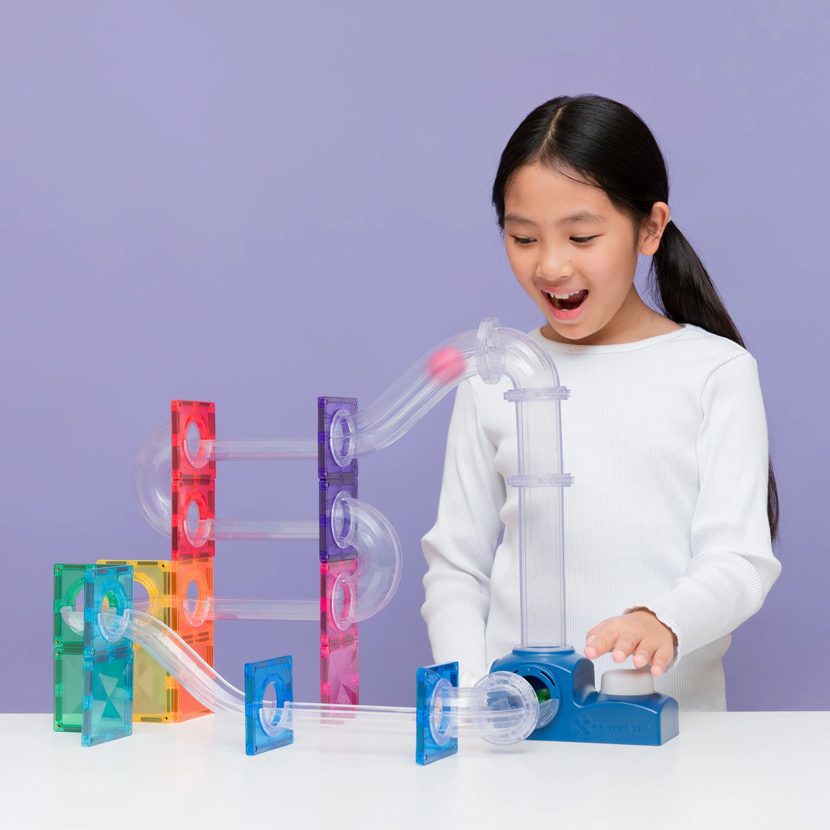 A young girl with dark hair in a ponytail, wearing a white long-sleeve shirt, is smiling and playing with a colorful transparent magnetic tile marble run toy set on a white table against a solid lavender background.