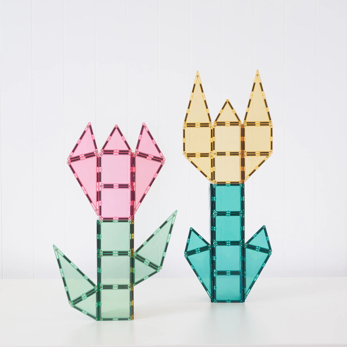 Two translucent flower-shaped structures made from colorful magnetic building tiles, one pink and green and one yellow and teal, displayed on a white surface against a white paneled background.