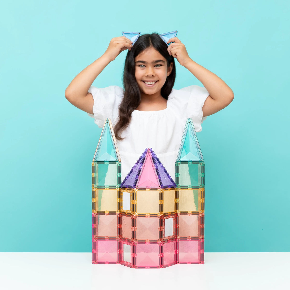 A young girl with long dark hair smiles while adjusting blue cat-ear magnetic tiles on her head, standing behind a colorful magnetic building block castle on a white table against a light blue background.