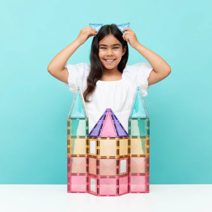 A young girl with long dark hair smiles while adjusting blue cat-ear magnetic tiles on her head, standing behind a colorful magnetic building block castle on a white table against a light blue background.