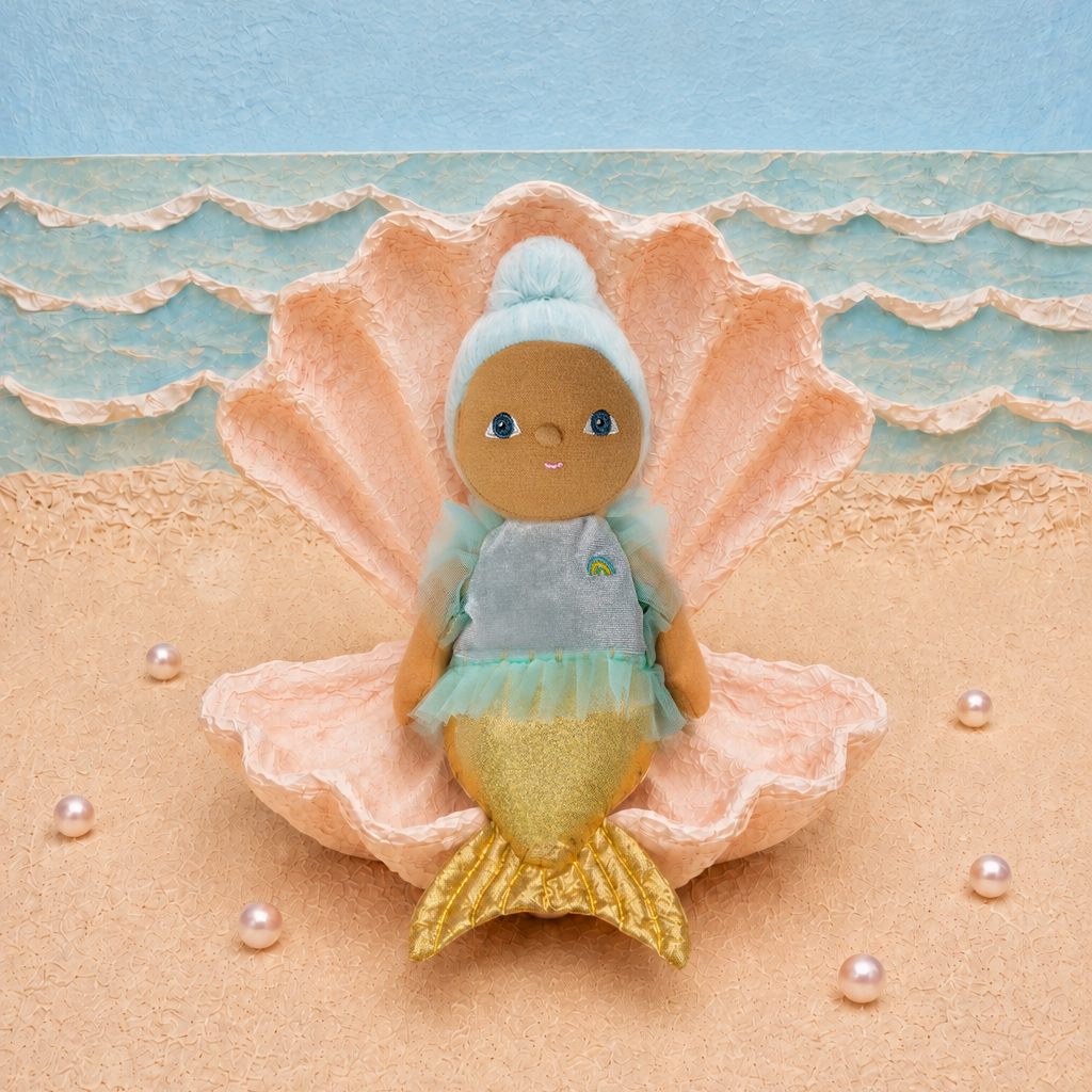 A small mermaid doll with blue hair, gray top, and gold tail sits inside a large peach seashell on a sandy beach-style backdrop with blue ocean waves and scattered pearls.