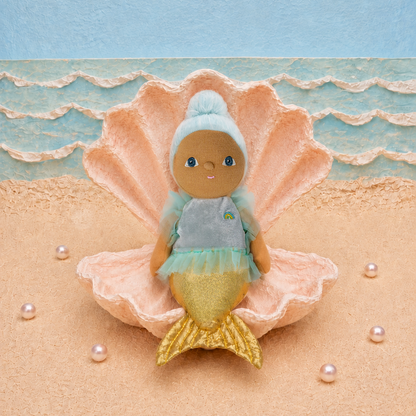A small mermaid doll with blue hair, gray top, and gold tail sits inside a large peach seashell on a sandy beach-style backdrop with blue ocean waves and scattered pearls.