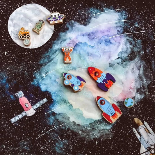 A flat-lay collection of colorful wooden toy space-themed cookies arranged on a Wonderie play cloth with a dark starry background and a painted nebula. The figures include rockets, a space station, a satellite, a UFO, a moon, and an astronaut.